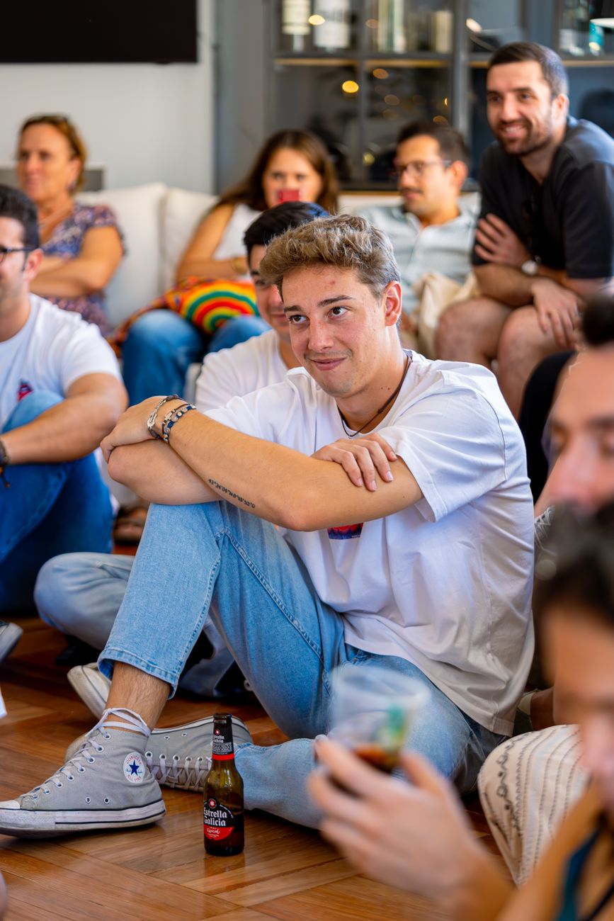 smiling young man sitting on floor with beer bottle listening among other people