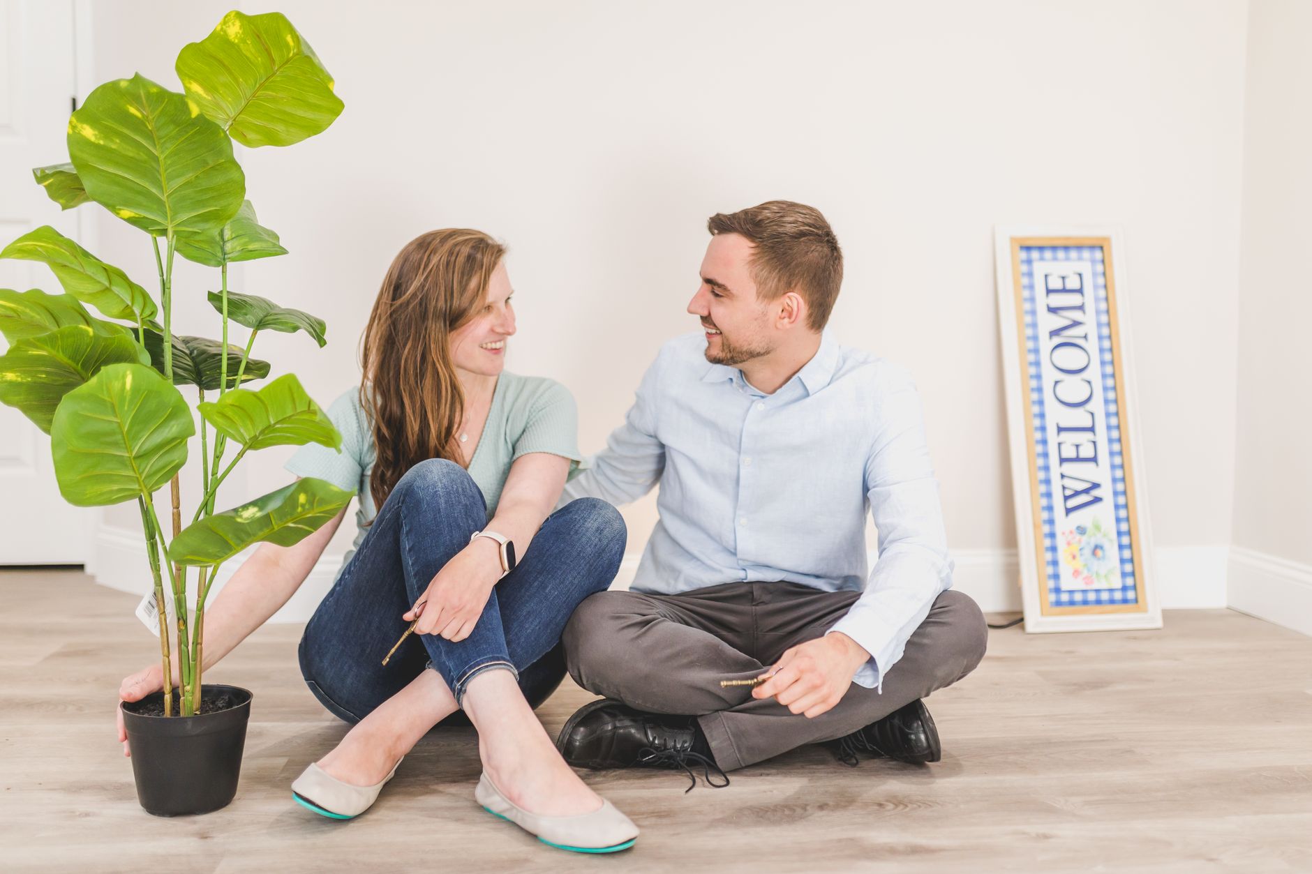 smiling couple sitting on wooden floor beside a houseplant and welcome sign