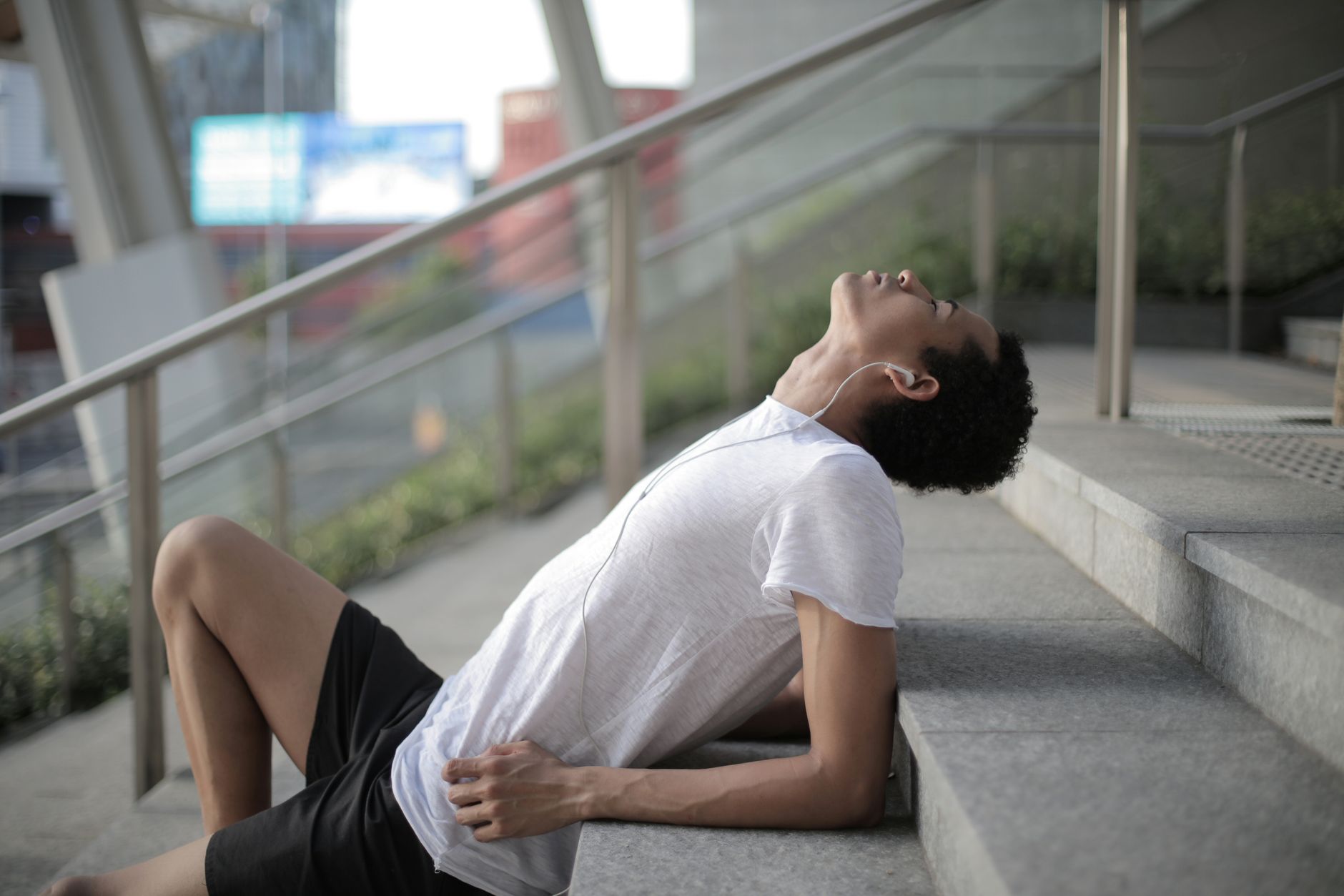 tired millennial ethnic male athlete listening to music and resting after training on stairs in city